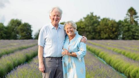 Ian Jamie owns the farm with his wife Pippa and the couple are seen here celebrating their 40th wedding anniversary with a glass of bubbly in a lavender field. Both are wearing light blue and they are standing together looking happy.