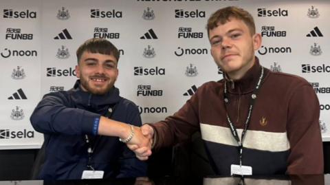 Kendrick Pardoe, wearing a navy jacket, shaking hands with a man wearing a burgundy quarter zip jumper. They are sitting behind a desk at Newcastle United.