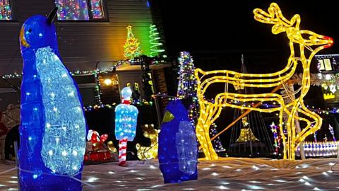 Illuminated penguins and a reindeer stand on a carpet of fake snow, surrounded by fairy lights. A house is in the background with lights in the window and trees on top of the porch.