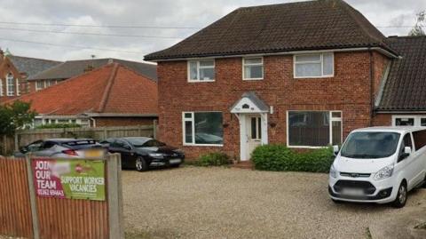 A detached two-storey house with white framed windows. There is a large gravel driveway with two cars and a van parked. 