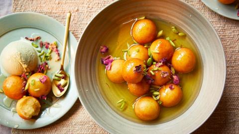 Gulab jamun on a plate with pistachio ice cream, saffron syrup and topped with dried rose petals and pistachio nibs.