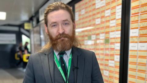 Elliott Badger with brown hair and a beard wearing a grey suit and green lanyard standing in Northampton railway station infront of a wall of bright orange train tickets