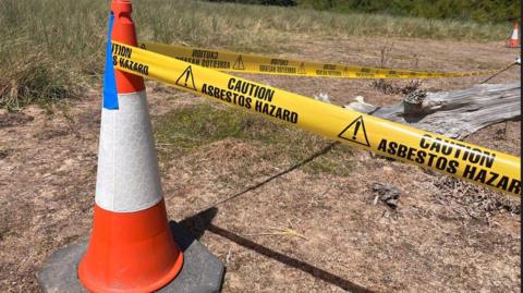 An orange and white cone is in the foreground with a yellow tape affixed to it, reading Caution Asbestos Hazard. It is by the side of parched grass and a dried out piece of tree trunk.