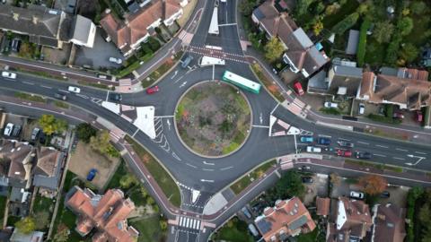 Aerial view of a roundabout. A red cycle lane is visible on roads to the left and right and on the roundabout itself. 