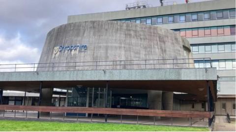 The front of Shropshire Council's headquarters. It is a large building with dozens of windows, with a circular dome in front of it. The blue and yellow Shropshire Council logo is on the dome. There is an area of grass and wooden fence in front of it.