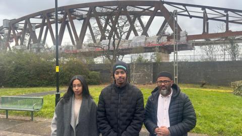 Three people - a woman and two men - stood in front of a HS2 viaduct. The woman has black hair and a grey coat. The man in the middle had a black hat and black coat and the man on the right has a black hat, black coat and white shirt. Behind them is a patch of grass then a large rust coloured structure