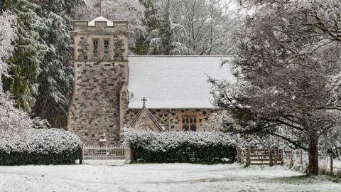 A church is covered in snow like the landscape around it. Trees surround the old building with branches dusted in the white stuff.