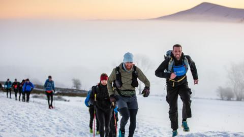 Racers run and trek through snow holding walking poles. They wear warm clothing. A mountain can be seen in background.