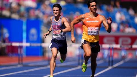 Jake is wearing the Great Britain kit of a blue vest with red writing on it and dark blue shorts. His name is on his vest. He is trailing behind Spain's Jesus David Delgado, who is wearing an orange top with 'Espana' across the chest.