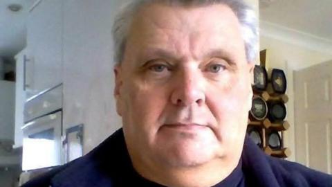 A head shot of a man in his home, he has grey hair and is wearing a blue top.
