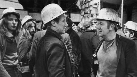 A black and white photograph of a group of men with hard hats on and in worn, dirty clothes. McGuinness stands on the right talking to another man. He has brown hair with large sideburns.