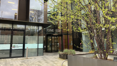 The outside of the West Northamptonshire Council offices at One Angel Square, Northampton. It is predominantly a glass building. On the right of the image is a thin tree coming out of a large planter.