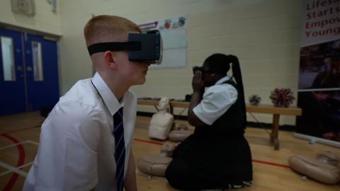 A male and female pupil wear a VR headset in a school gym hall.