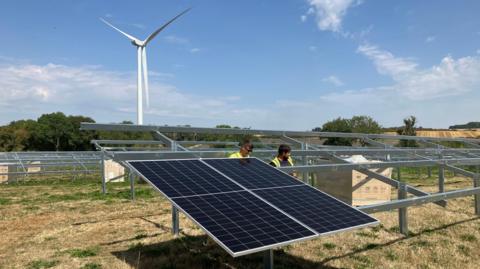 A picture of two men installing solar panels. They are both wearing yellow high visibility jackets and there is a wind turbine behind them and it is a sunny day with blue skies. 