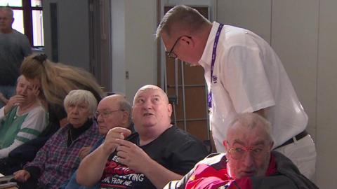 A man in a white short-sleeved shirt stands above another man who is sitting down and wearing a black t-shirt while the pair have a chat in a room full of people.