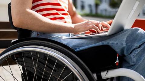 A close-up image of a wheelchair as a woman sits in and works on a laptop. Her jeans and red and white T-shirt and hands and forearms are visible.