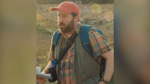 Man wears orange hat, checked shirt and rucksack, and walks along outside