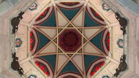 Ceiling of a church, taken from the floor below, showing an octagon style shape with a star shape formed in stained glass, coloured in blue, red, white, and gold