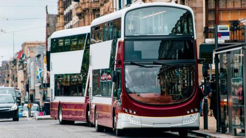 Two double-decker Lothian Buses are parked at a bus stop in a busy road. They are maroon and white.