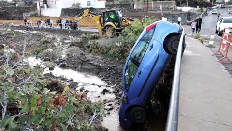 A blue car is nose down in a ditch by a bridge. A tractor is removing debris and mud and people are watching nearby.