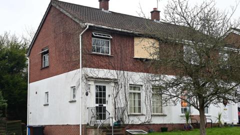 A semi-detached house with red and white brick. One of the upper windows is boarded up and other upstairs windows are open.