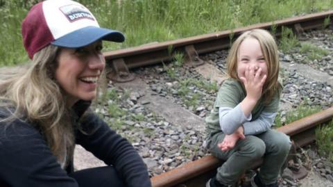 Nadia McSheffrey, a woman with blonde hair wearing a cap with a blue rim, sitting on a disused railway line with her young son - aged about five - who is laughing and covering his mouth with his hand.