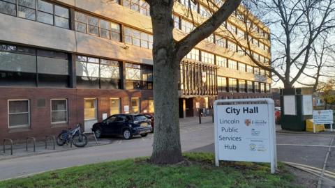 This photo shows a Lincoln City Hall building with multiple windows. In front, there's a white sign reading "City Hall, Lincoln Public Services Hub," along with logos and details about its services. A tree is in the foreground, and parked cars and bicycles can be seen near the entrance.