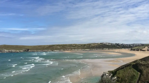 The picture shows a wide view across Crantock Beach near Newquay, looking over the sweeping curve of sand at the mouth of the River Gannel. In the foreground, pale green‑blue seawater rolls in with small, broken waves, creating streaks of white surf that angle across the bay. 