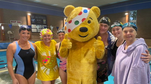 Six members of the BBC Cornwall team in swim wear lined up in front of a swimming pool with Pudsey bear stood in the middle of the group