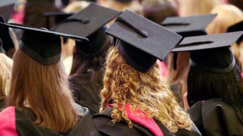 The image shows a graduation ceremony viewed from behind the audience of graduates. Several people are sitting closely together, all wearing black graduation gowns and square black caps with tassels hanging from them.
One graduate has long, curly blonde hair spilling down the back of their gown, with a bright red sash draped over their shoulders.