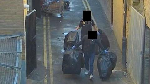 CCTV of two people carrying black bin bags down a back alley. Their faces have been obscured.