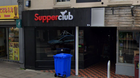 A general view of the exterior to SupperClub. The club's entrance is black with the name of the bar written above. A blue rubbish bin sits outside the entrance. 