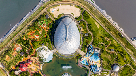 A drone photo from above Oriental Eden. The biome is shaped like an egg and it is surrounded by gardens and a small lake with fountains. There is a larger lake surrounding the site. The lake coastline echoes the shape of the top of the biome and there is a road inset from the shoreline.
