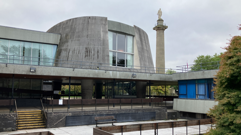 Large building that is made of grey concrete with rows of dozens of windows. There is a large funnel-shaped structure above an entryway. In the background is Lord Hill's Column, a tall doric column in light coloured stone, topped with a statue