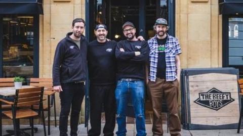 Four men standing outside a storefront built in honey-coloured stone. They are all wearing caps and three have branded restaurant tops on. The fourth is in a checked shirt. Next to them, a sign reads "The Beefy Boys"