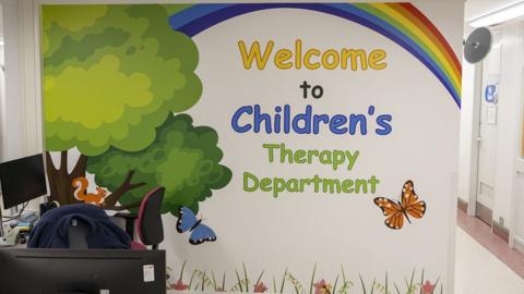 The reception area at the new unit with a desk and computer and a wall with a mural of trees, flowers, butterflies and a squirrel with a rainbow in the sky.
The words Welcome to Children's Therapy Department are painted beneath the rainbow.