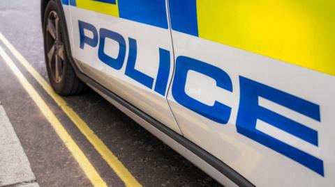 The sider of a car marked in large blue letters with the word POLICE as well as blue and yellow markings. The car is parked on double yellow lines.