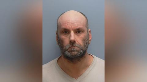 Elliot Jones is looking at the camera in a police custody photo. He is balding and has a greying beard and moustache. He is wearing a cream pullover.
