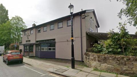 Google streetview of two-storied cream-coloured bar with a traditional lamplight outside and car parked on roa. Trees line both sides.