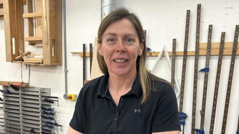 Tracy Burns - a woman with shoulder length hair wearing a black polo shirt looks smiling into the camera. She is standing at a wooden work bench with tools on it with other construction tools and wood on the wall behind her