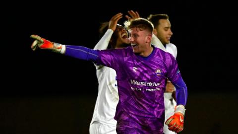 Braintree goalkeeper Mason Terry, in a purple shirt and orange gloves, points at teammates to celebrate scoring a stoppage time goal against Truro in January.