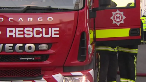 The front of a red fire truck, with two fire officers behind the door which has the Devon & Somerset Fire & Rescue Service emblem on it.