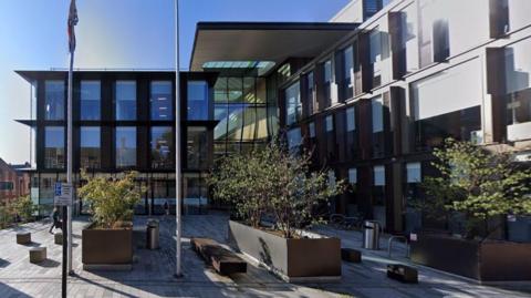 Glass and steel-fronted building three-storey office building with small trees and seats outside. There is a paved piazza with flagpoles.