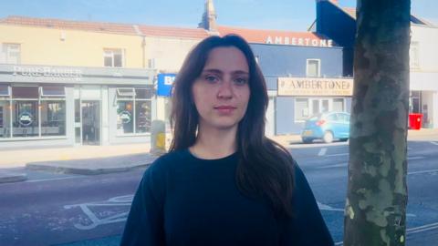 A young woman with dark hair and a navy jumper is stood on Fishponds Road where the accident happened. It is a busy road with lots of businesses.