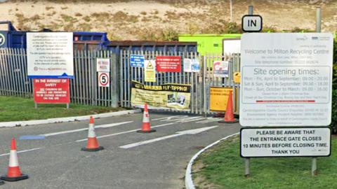 The outside of a closed Milton Recycling Centre, with the gates shut and traffic cones placed on the driveway. There is a sign showing its opening hours on the right.