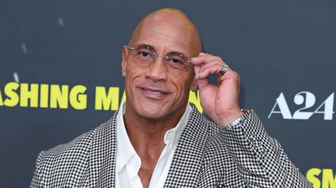 Dwayne Johnson raises his hand to his glasses while standing on the red carpet for his latest film The Smashing Machine