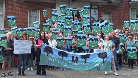 Approximately 40 people are standing on steps outside Swindon council civic offices holding blue and green posters that read "Keep our old golf course green".