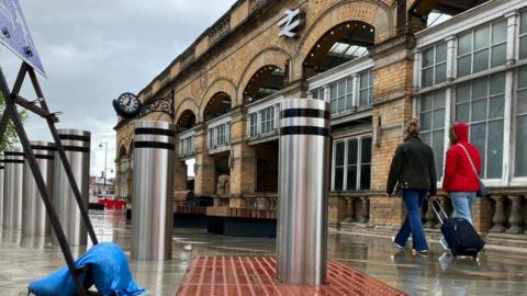 A bollard rises out of red tactile paving, with York Railway Station in the background.