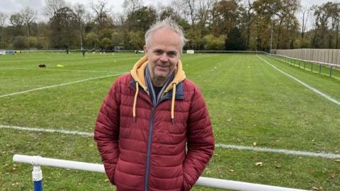 Paul Rushton poses at the ground of Newport County AFC, leaning against a metal barrier with the pitch in the background. He has grey hair and is wearing a red padded jacket over a yellow hoodie. Trees can be seen in the background beyond the pitch