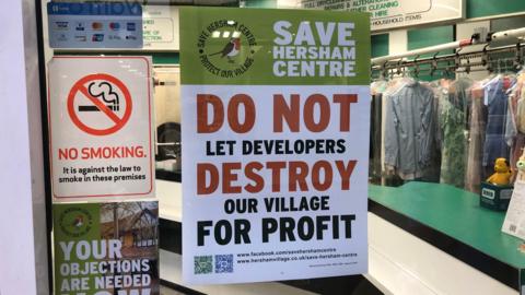 Poster from campaign group Save Hersham Centre in the window of shop. The green and white poster has red, black and white writing and says: "Do not let developers destroy our village for profit."